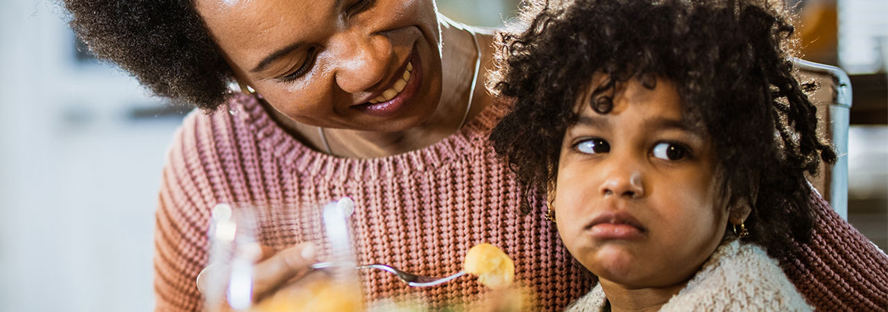 Happy single black mother trying to feed her small girl who is sulking.