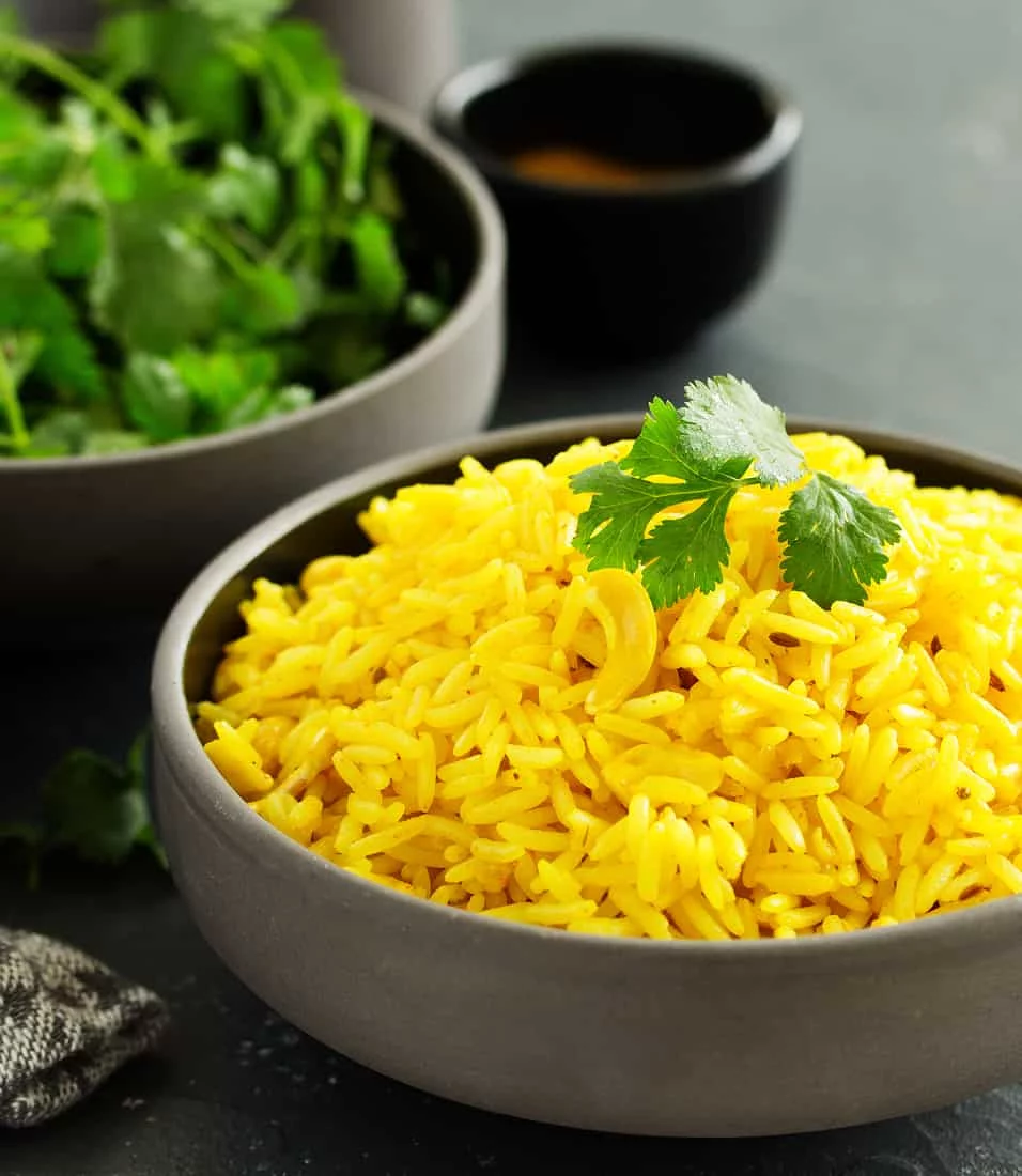 A close-up of vibrant yellow rice served in a gray bowl, garnished with fresh cilantro leaves. The setting includes additional bowls of herbs and sauce in the background, creating a rustic and appetizing presentation. The dark surface contrasts with the bright colors of the food.