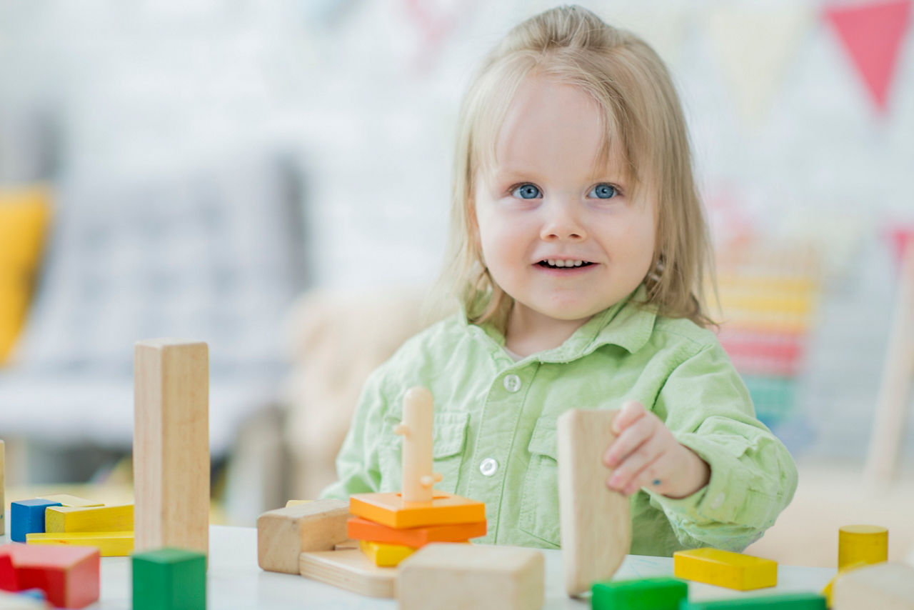 A Caucasian female toddler with blonde hair is indoors in a daycare. She is wearing casual clothing. She is looking toward the camera while building with toy blocks.