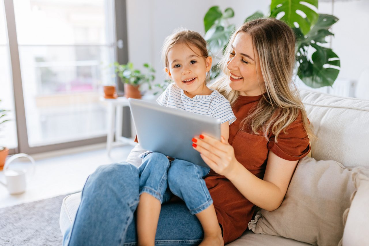 Eine Frau sitzt mit einem lachenden Mädchen auf dem Schoß auf dem Sofa, beide schauen gemeinsam auf ein Tablet.