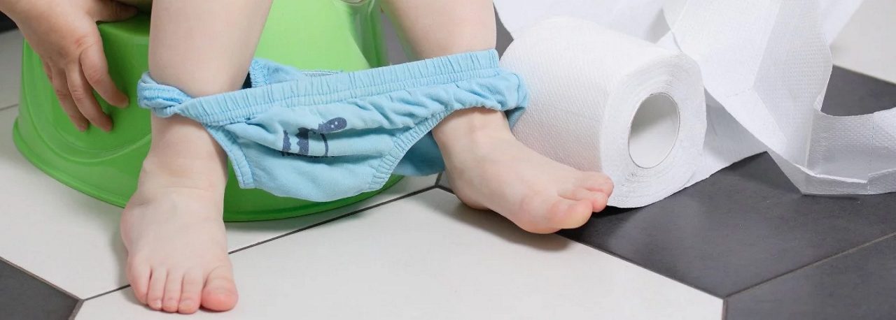 Ein Kleinkind sitzt auf einem grünen Töpfchen, trägt blaue Unterhose und neben ihm liegt eine Rolle Toilettenpapier auf Fliesen.