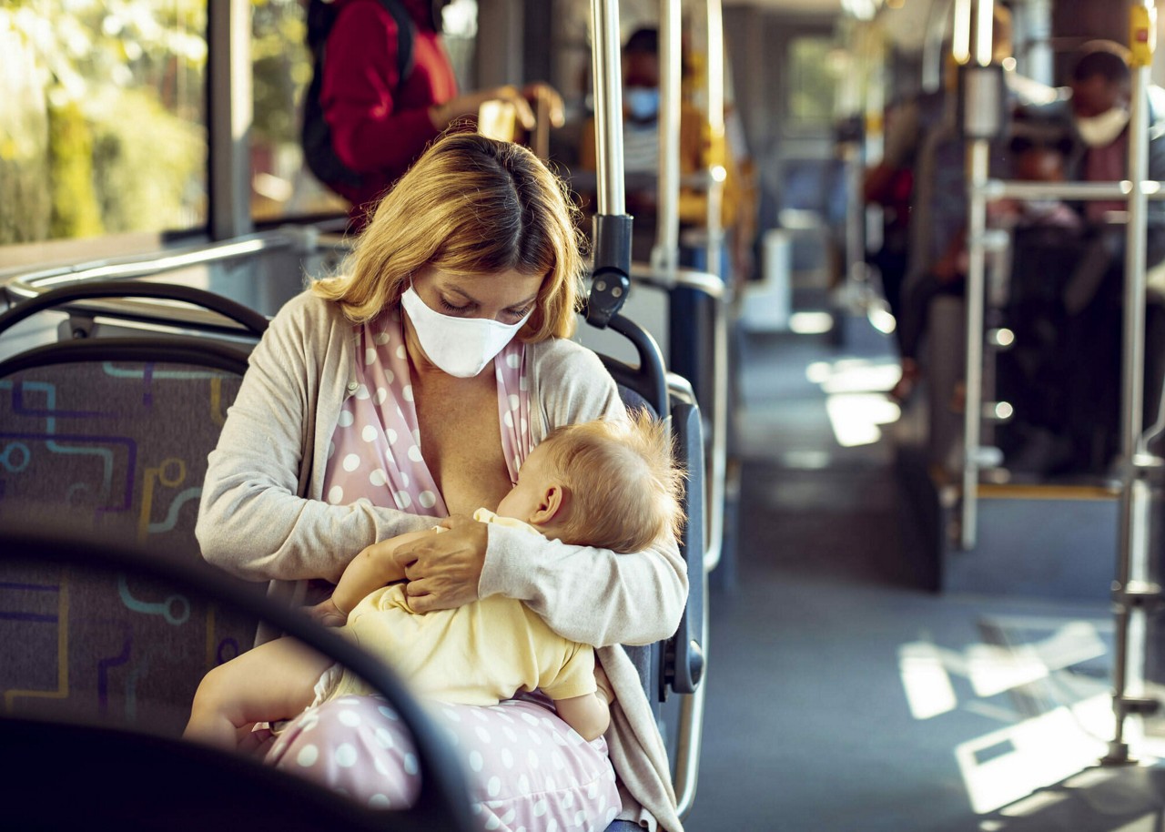 On voit une femme allaiter son bebe assise dans un bus public. Le decor se caracterise par un interieur bien eclaire avec des passagers en arriere-plan. Le bebe est vetu d une tenue jaune clair, et la femme porte un haut a pois rose avec un cardigan leger. La scene capture un moment de soins maternels dans un environnement de transport en commun.