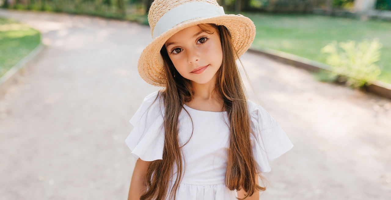 Little fashionable lady with long eyelashes looking with interest while standing on the road in elegant hat. Outdoor portrait of shy brown-haired girl wearing cute white gown., Little fashionable lady with long eyelashes looking with interest while standing on the road in elegant hat. Outdoor portrait of shy brown-haired girl wearing cute white gown.
