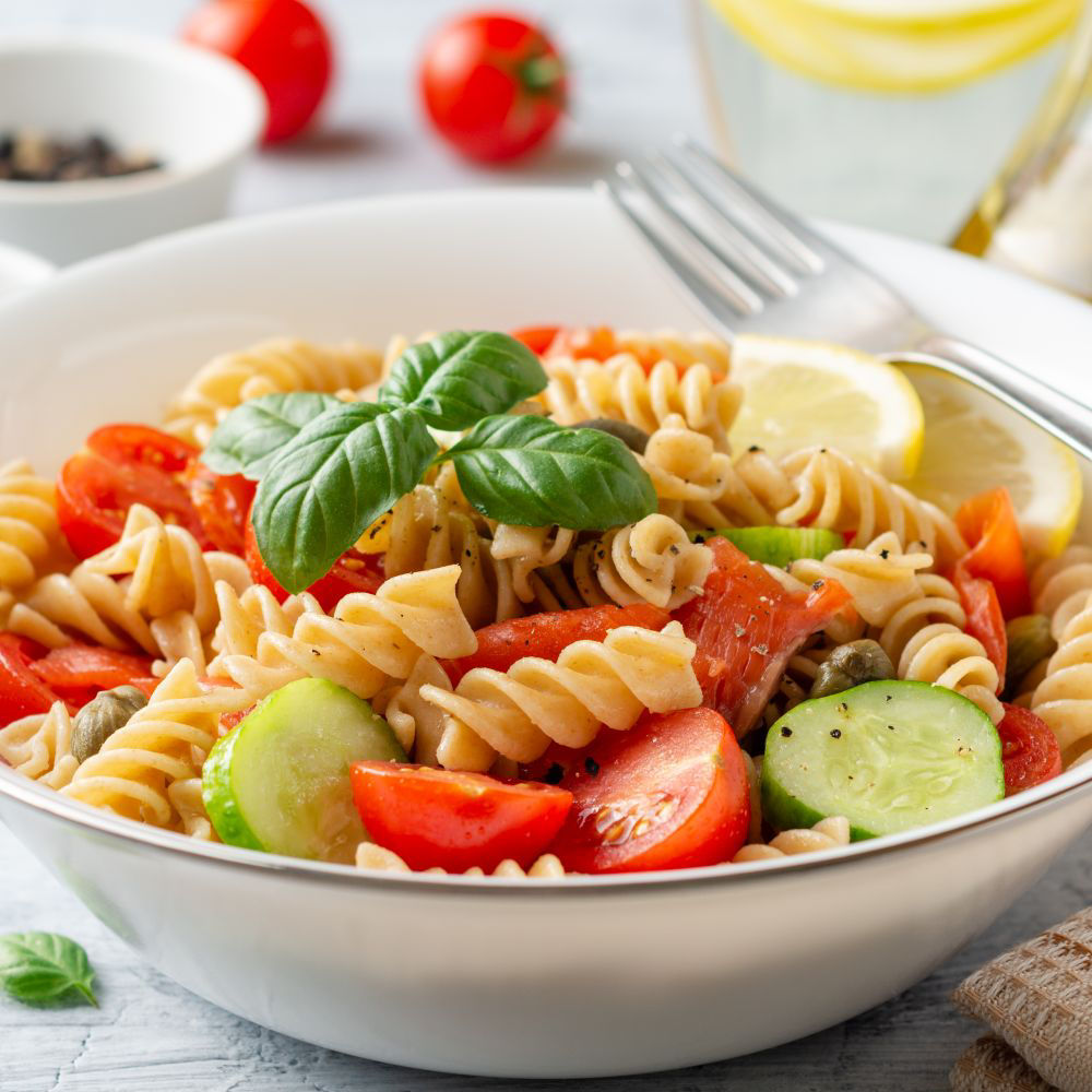 Whole wheat pasta salad with cucumbers, cherry tomatoes, salted salmon and capers on concrete background. Selective focus.