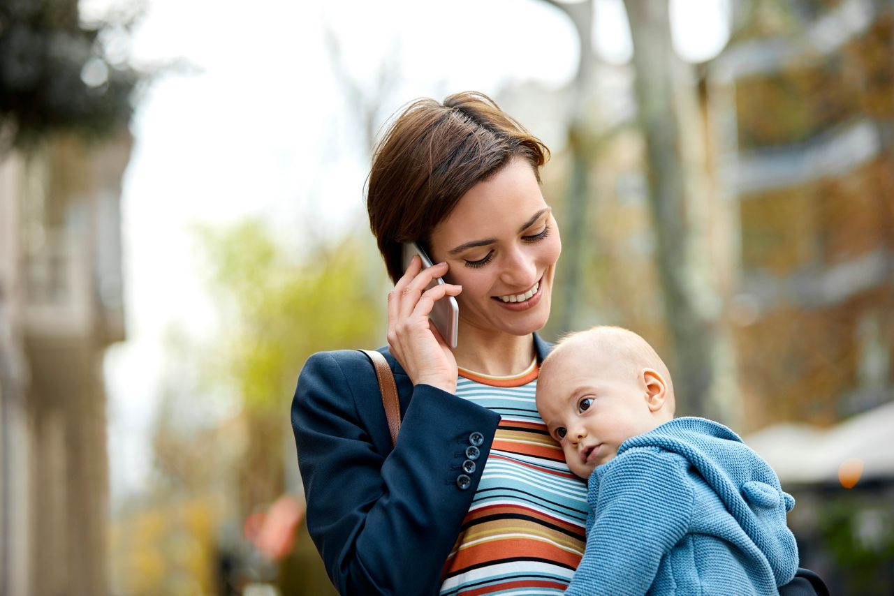 Mid adult mother talking on mobile phone while carrying son. Smiling woman is using smartphone while looking at baby boy on street. They are in city.