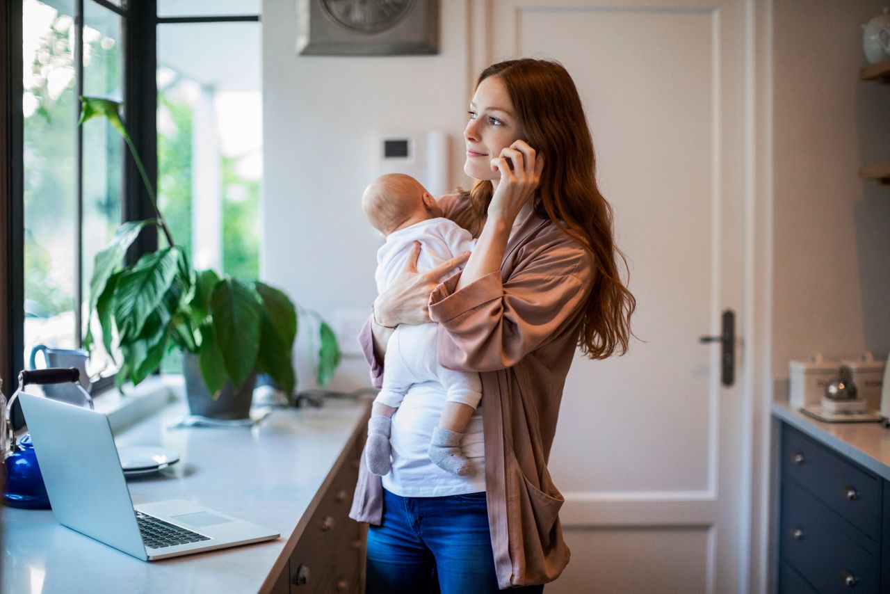 Happy mother looking away while carrying baby boy talking on smart phone in kitchen. Smiling redhead woman is standing with newborn son by laptop on kitchen counter. They are at home.