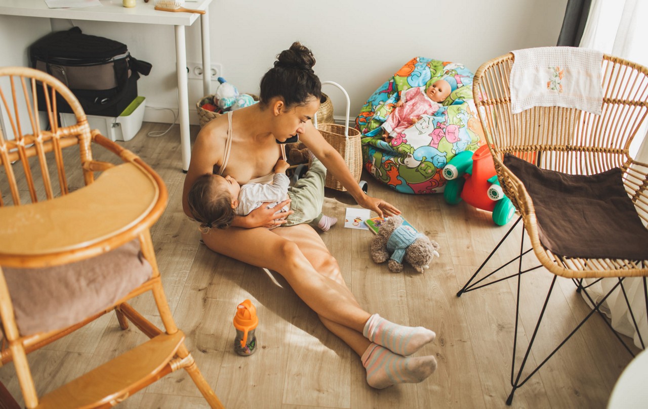 Une femme est assise sur le sol tenant un bebe dans une salle de jeux lumineuse et confortable. Le cadre comprend des meubles en bois, des jouets colores et un biberon visible a proximite. La piece a une esthetique chaleureuse et naturelle avec un eclairage doux et un decor ludique.