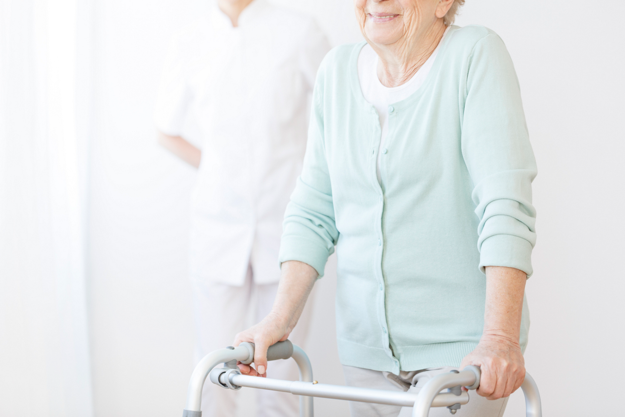 Eine ältere Frau in mintgrüner Strickjacke nutzt einen Rollator, im Hintergrund steht eine Pflegekraft in Weiß.