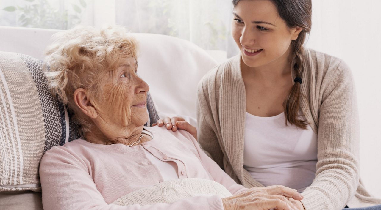 Eine junge Frau sitzt neben einer älteren Dame auf dem Sofa und hält ihre Hand, beide lächeln einander freundlich an.