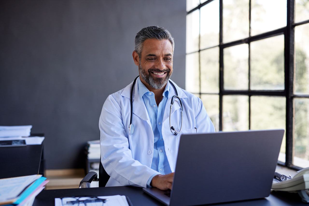 Mid adult doctor smiling while having online consultation on his laptop. Healthcare middle aged man conducting online consultation in an office setting. Indian doctor in conversation with patient over video conferencing using computer.