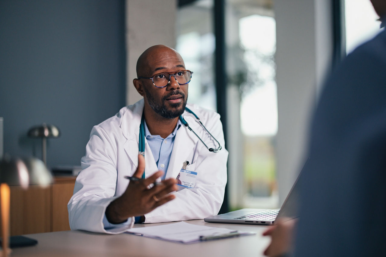 A doctor in a white coat speaks with a colleague across a desk, gesturing with hands in a medical office. Stethoscope hangs around neck; laptop and papers on the desk emphasize clinical consultation.