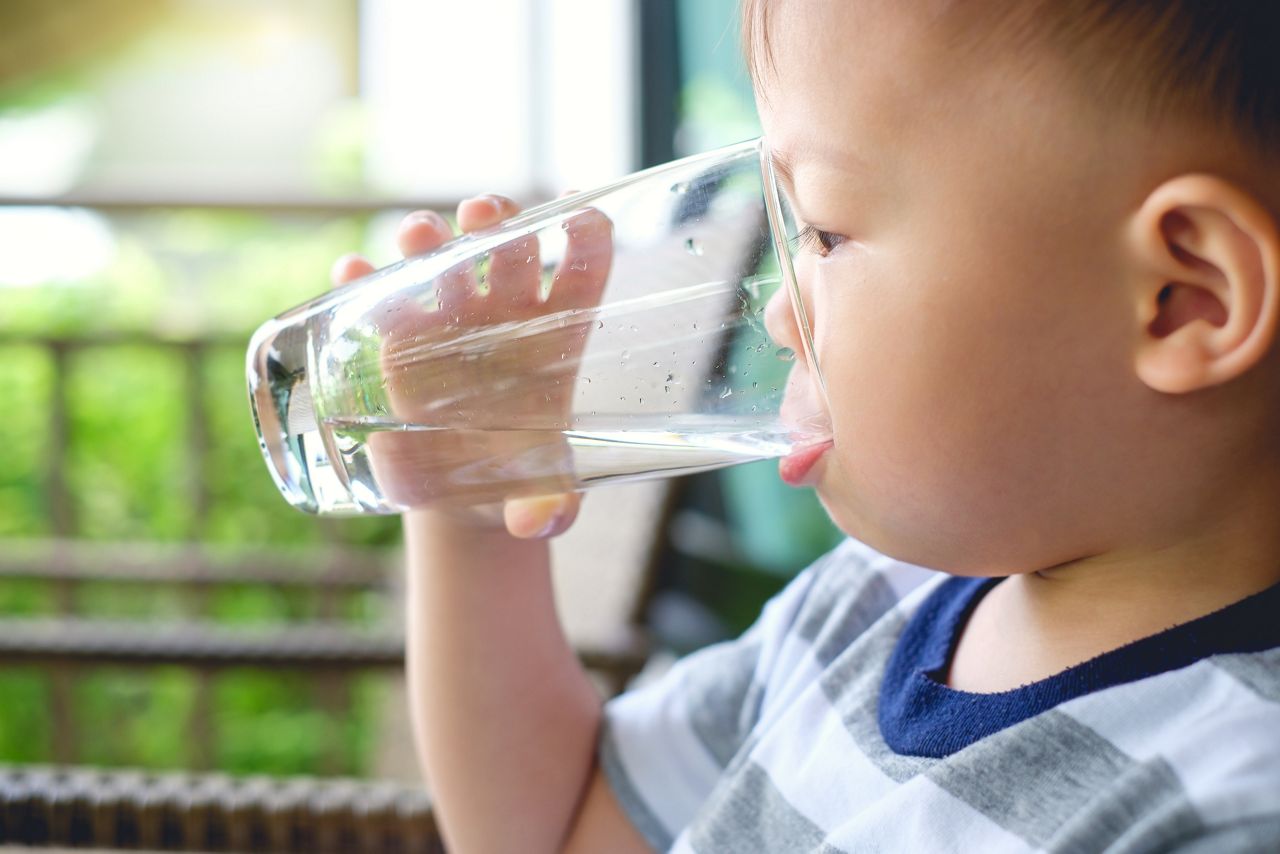 Cute thirsty little Asian 2 years old toddler baby boy child holding and drinking glass of water by himself against green background near home garden, best beverages for child's health concept