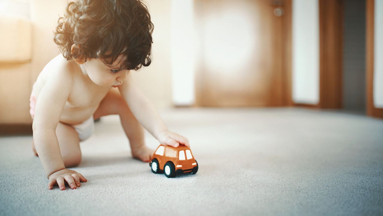 Closeup of a two year old toddler with curly brown hair sitting on the living room floor and playing with an orange toy car.He's wearing diapers only.