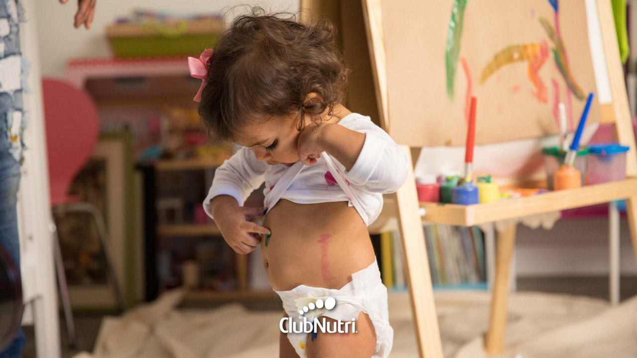 Niña jugando con pintura apuntando a su barriga con el dedo pintado