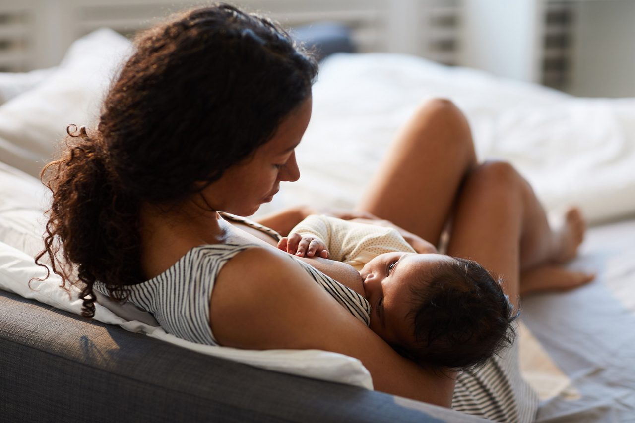 Peaceful loving youngmother sitting on bed and leaning on headboard while feeding baby with breast