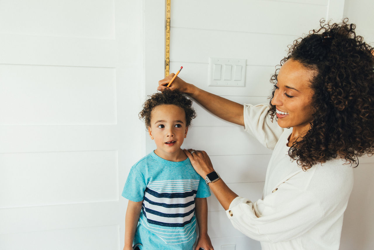 A mom measures her son against a ruler and wall to see how tall he is inside their home. The boy is growing fast at 4 years old and is excited to grow and learn with his mom teaching him.