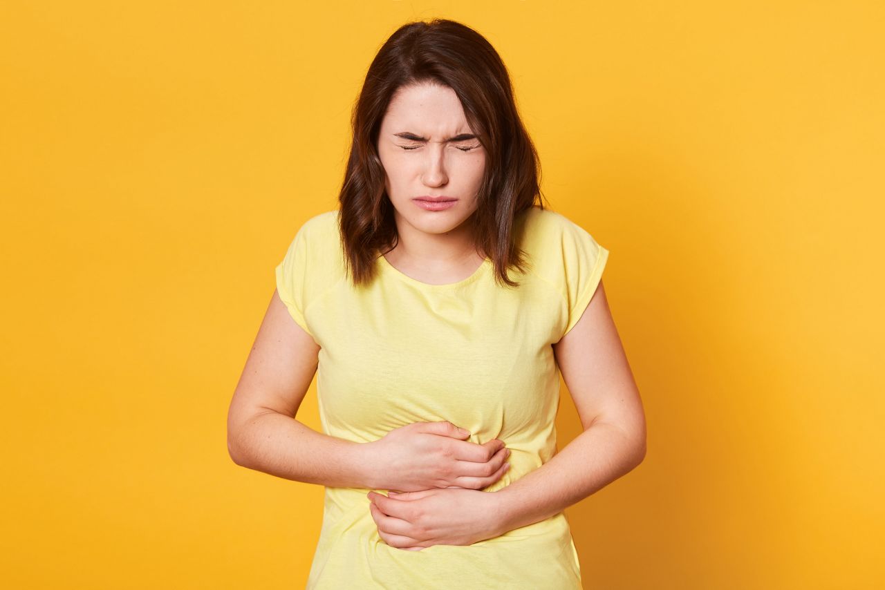 Close up portrait of young Caucasian woman with awful stomachache on yellow background, eats something expired, has intoxication, stands with closed eyes and keeps hamd on belly. Health concept.