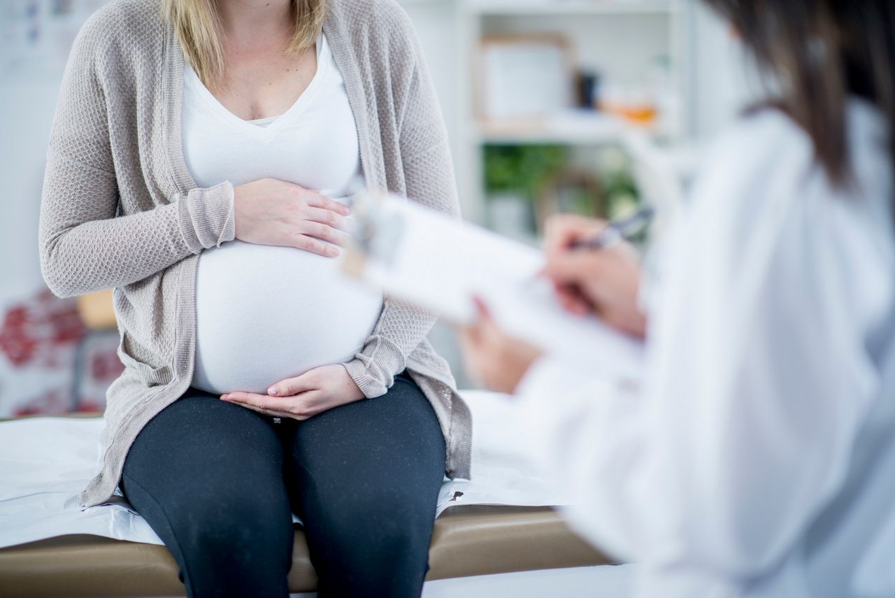 A pregnant woman and her doctor are indoors in a medical center. The woman is holding her stomach while the doctor writes information on a clipboard.