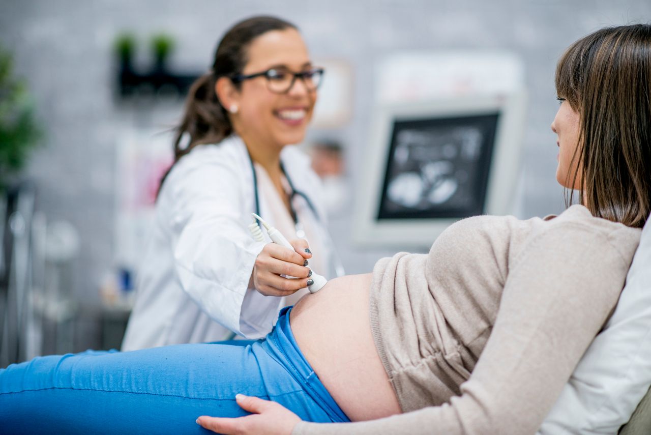 A pregnant Caucasian woman is indoors in a medical clinic. She is lying down while having an ultrasound. Her doctor is smiling with her while performing the procedure.