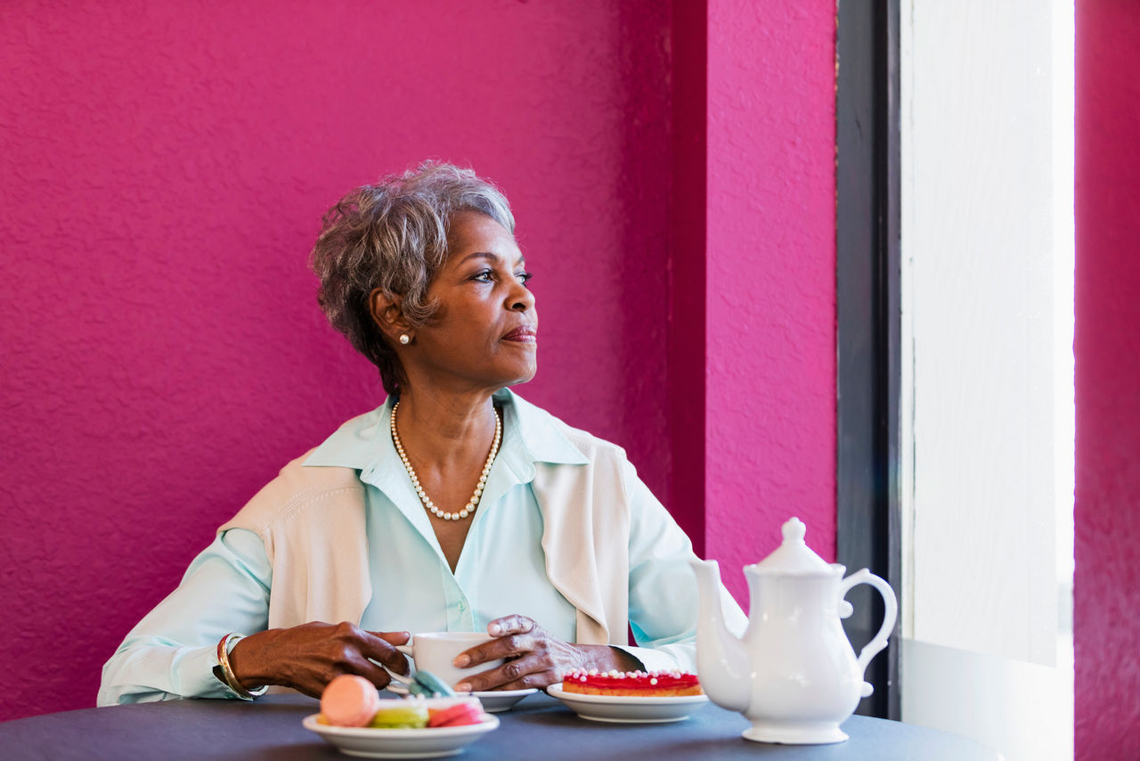 A senior African-American woman in her 70s sitting at a table indoors, drinking coffee, looking out the window with a serious expression.