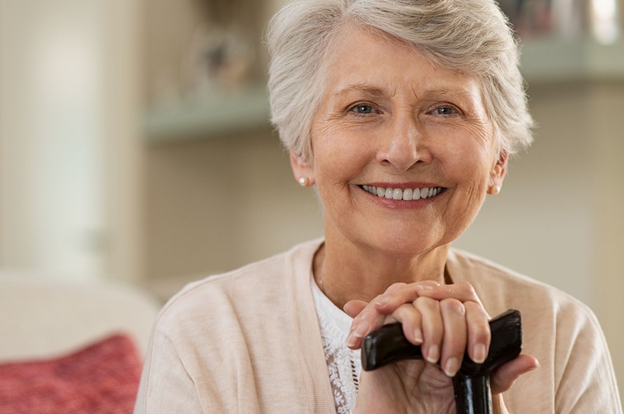 Retired woman with her wooden walking stick at home. Happy senior woman relaxing at home holding cane and looking at camera. Smiling grandmother sitting on couch.