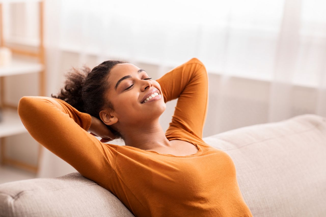 Enjoying Weekend. Happy Afro Girl Relaxing Sitting On Couch Holding Hands Behind Head At Home