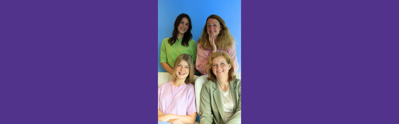 Drie glimlachende vrouwen zitten voor grote plant in modern kantoor.