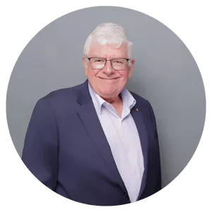 An older man is dressed in a navy blue suit and light-colored shirt, standing against a plain gray background. The setting is neutral and professional, with no visible text or logos. The image conveys a formal and composed mood.