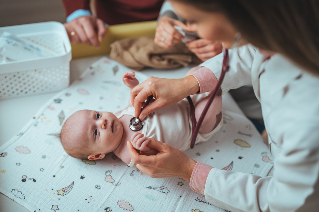 A Caucasian female pediatrician gently examines a newborn with a stethoscope. The baby rests on a changing pad, surrounded by medical tools, reflecting routine health check-ups in a clinical environment.