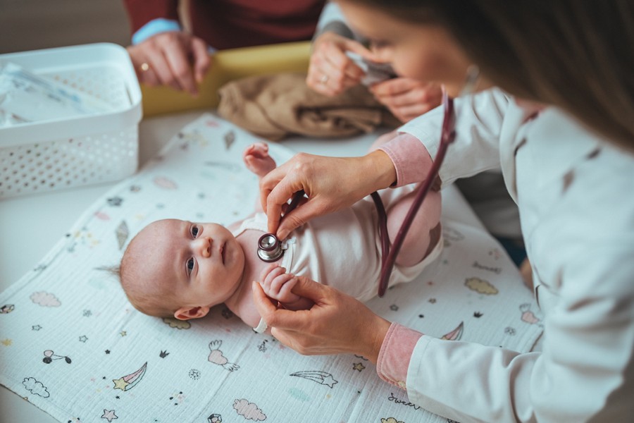 A Caucasian female pediatrician gently examines a newborn with a stethoscope. The baby rests on a changing pad, surrounded by medical tools, reflecting routine health check-ups in a clinical environment.