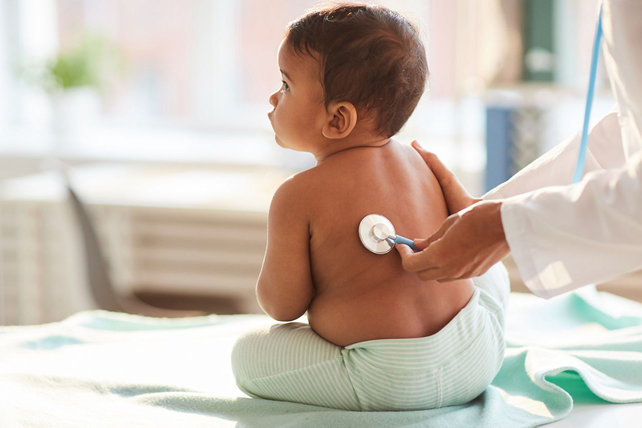Rear view of shirtless toddler sitting on the table while doctor listening to him with stethoscope