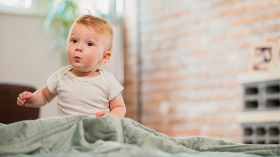 Ein Baby mit hellem Body sitzt auf einem Bett vor einer Ziegelwand, im Hintergrund steht eine Pflanze am Fenster.
