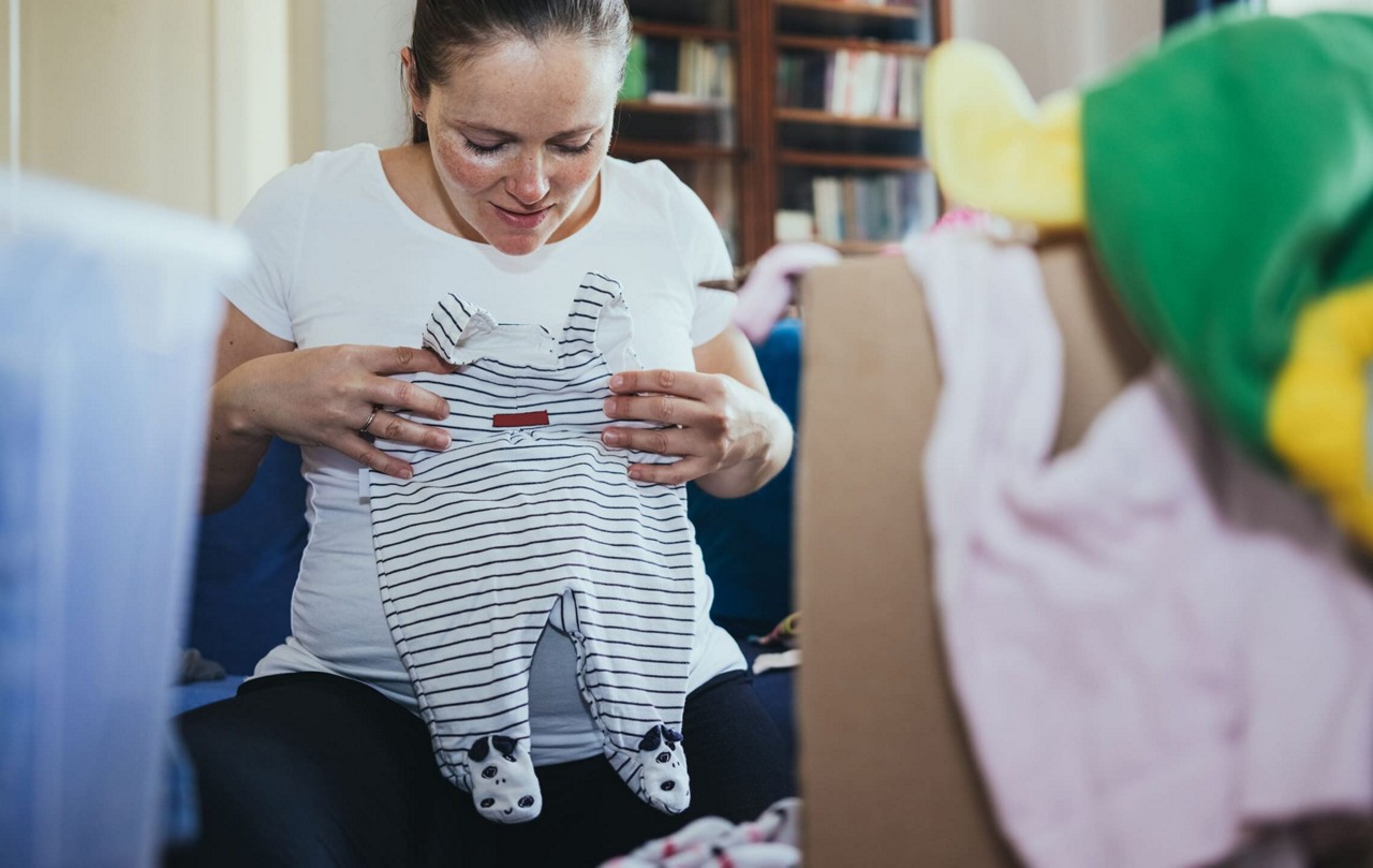 Une personne tient une grenouillere pour bebe rayee avec des details d empreintes de pattes sur les pieds. Le cadre semble etre un espace interieur confortable avec des etageres et des vetements de bebe eparpilles en arriere-plan. La scene suggere une preparation ou une organisation pour un bebe. La grenouillere comporte une petite etiquette visible mais pas de texte ou de logo clairement identifiable.