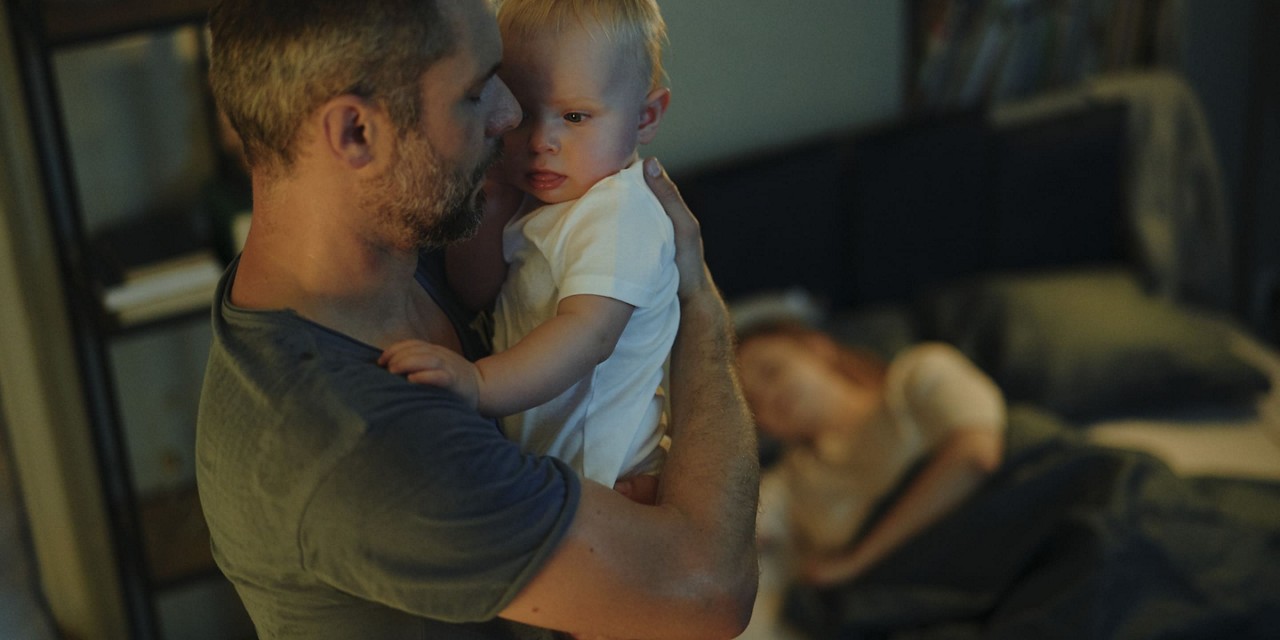 Un homme tient un bebe dans une chambre chaudement eclairee. Le cadre comprend un lit avec une femme reposant en arriere-plan, creant un moment familial serein et intime. La scene transmet un sentiment de soin et de tendresse, avec un eclairage doux et des tons sourds rehaussant l atmosphere chaleureuse.