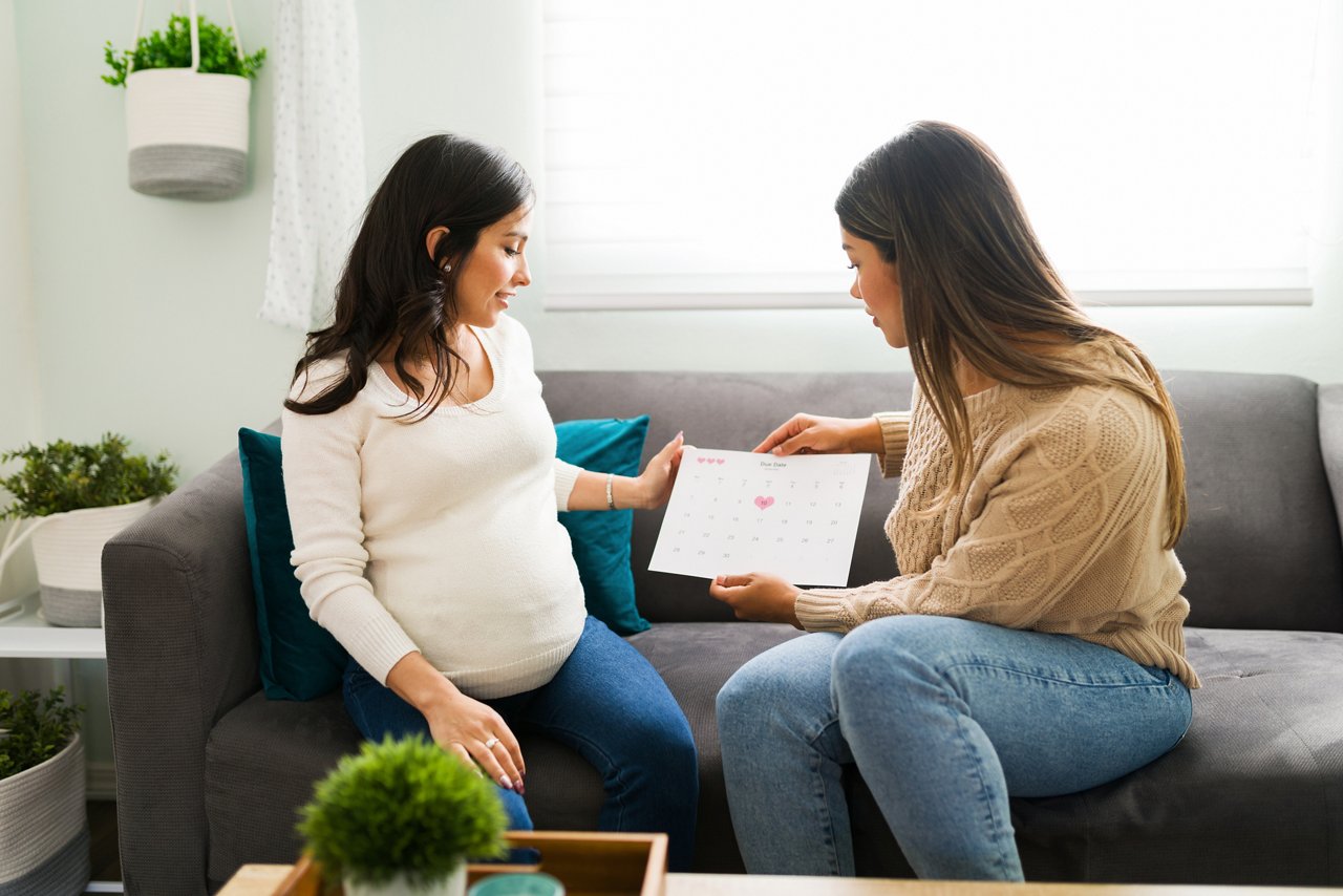 Beautiful pregnant woman and latin midwife holding a calendar. Doula and expectant mother talking about the due date while sitting on the couch