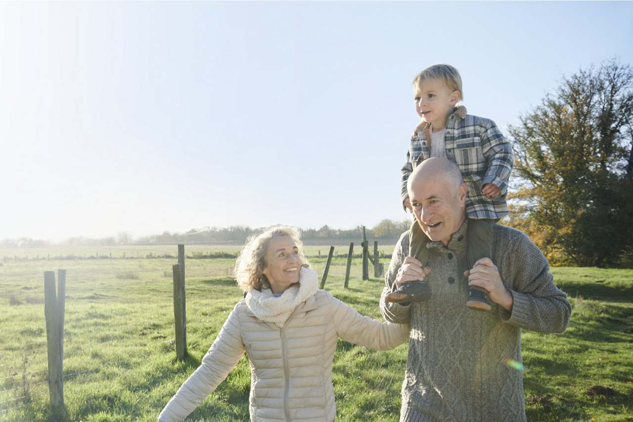 Oud echtpaar loopt met kind op schouders man over grasveld, op achtergrond bomen.