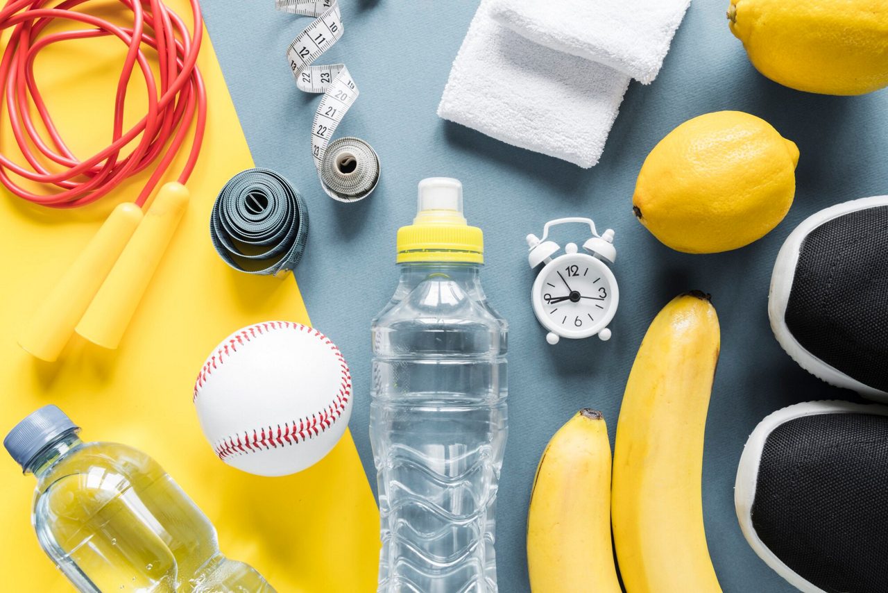 Platliggende opname van fitness- en wellnessbenodigdheden, waaronder een springtouw, meetlint, handdoek, waterfles, honkbal, wekker, bananen, citroenen en sportschoenen.