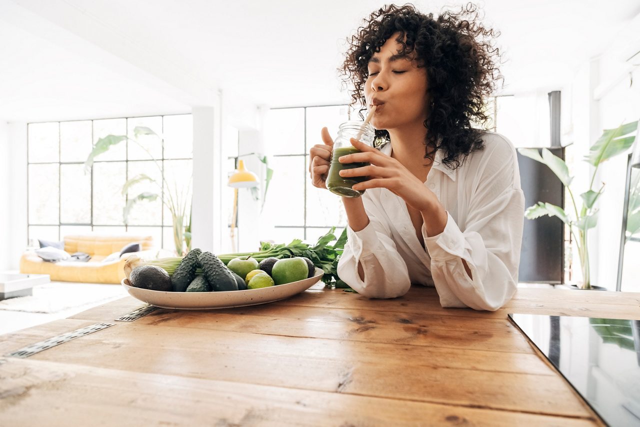 Afro-Amerikaanse vrouw die lacht terwijl ze een glas groen sap drinkt, genietend van een gezonde levensstijl.