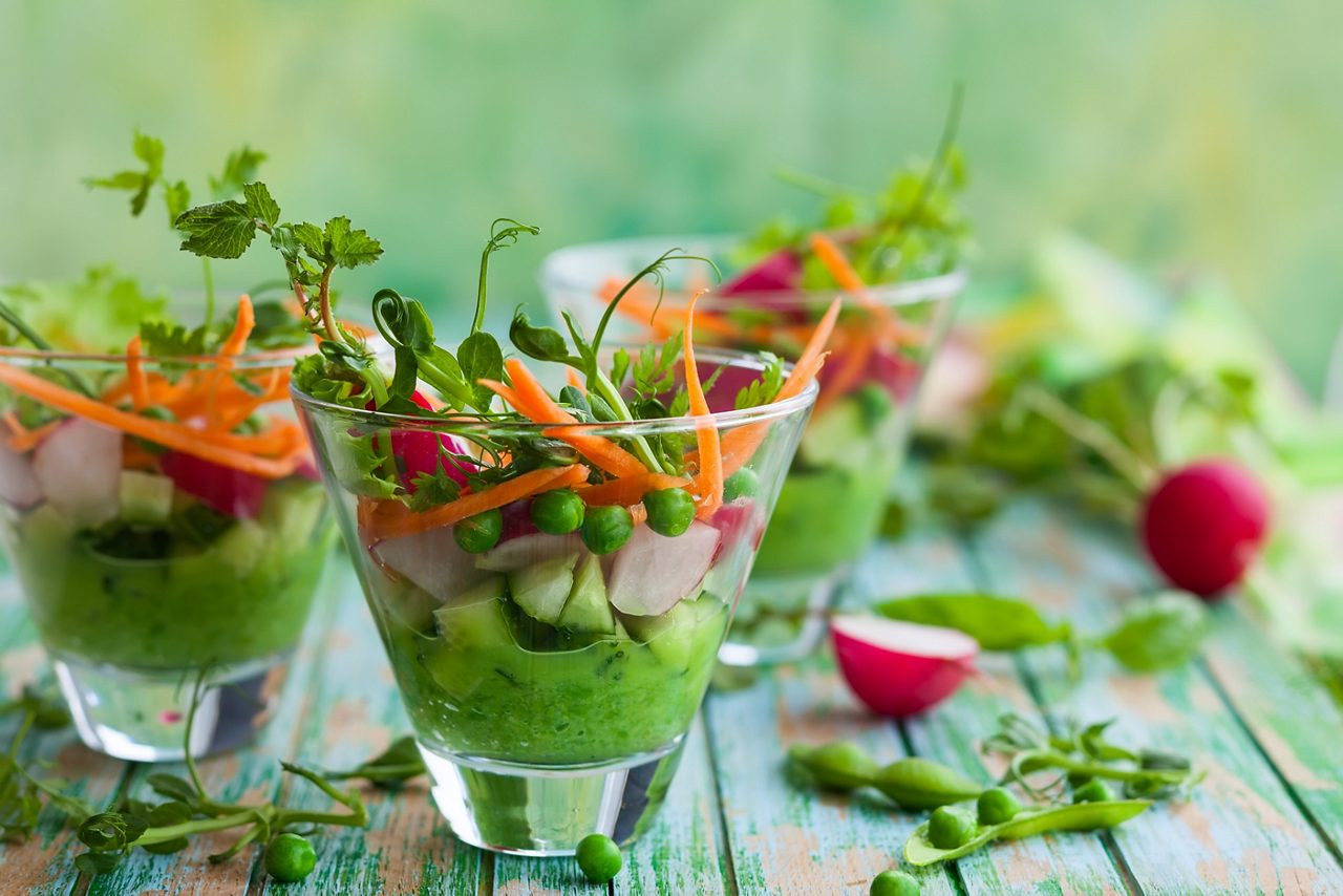Kleurrijke gelaagde groentesalades in glazen bekers met erwten, wortelen en bladgroenten.