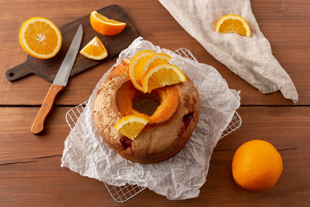 Bundt cake gegarneerd met verse sinaasappelschijfjes, omgeven door sinaasappelpartjes op een houten tafel.