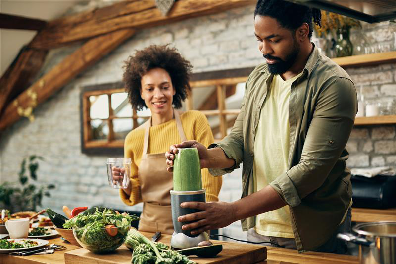 Glimlachend koppel in een keuken die een smoothie bereidt met behulp van een blender, omringd door groenten.