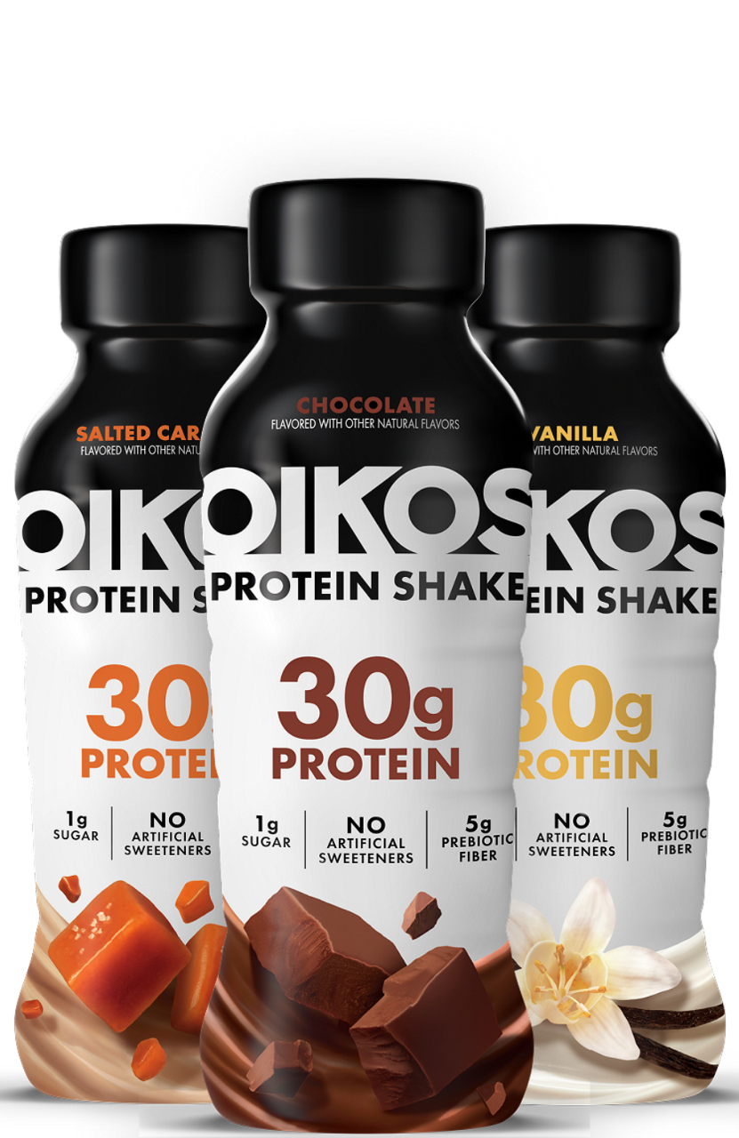 Three Oikos protein shakes are displayed in salted caramel, chocolate, and vanilla flavors. Each bottle prominently highlights '30g Protein' along with '1g Sugar', 'No Artificial Sweeteners', and '5g Prebiotic Fiber'. The bottles are visually appealing with flavor-specific imagery such as caramel cubes, chocolate chunks, and vanilla flowers.