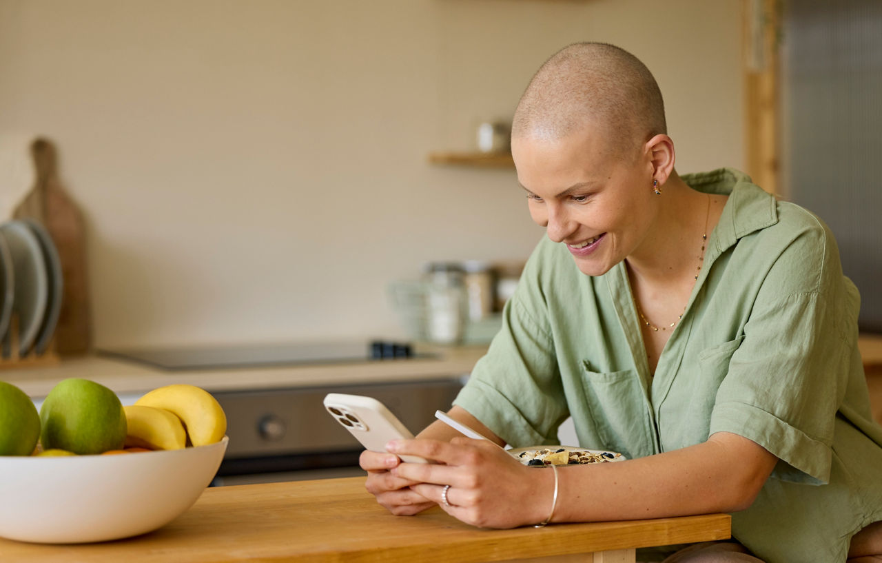 Smiling woman using smart phone while having breakfast. Female with shaved head is sitting at table in kitchen. She is at home.