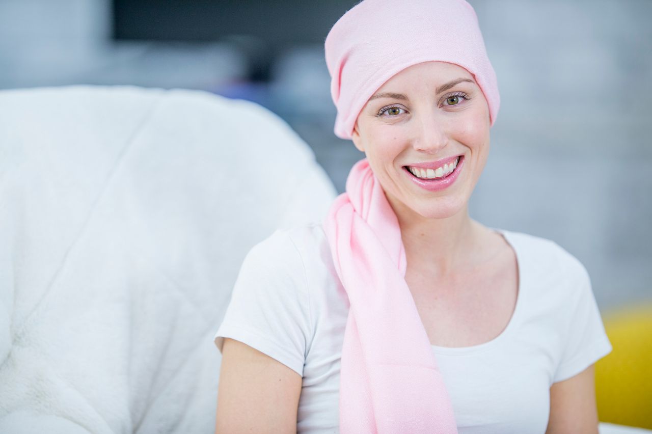 A Caucasian woman is indoors in her house. She has cancer, and she is wearing a head scarf to hide her hair loss. She is sitting in a chair and smiling at the camera.