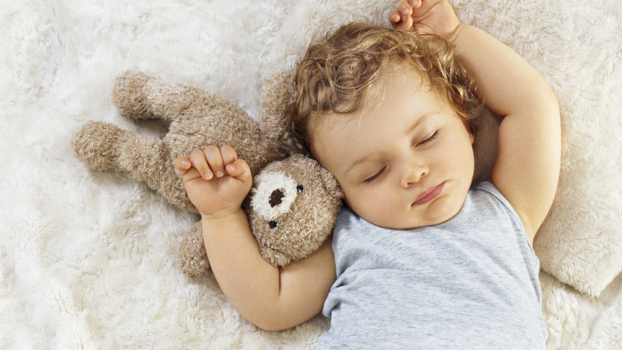 Portrait of a sleeping baby boy 11 months old lying down arms raised up, on a white synthetic fur blanket with a Teddy bear touching his head, his top raised showing his navel, view from above, France