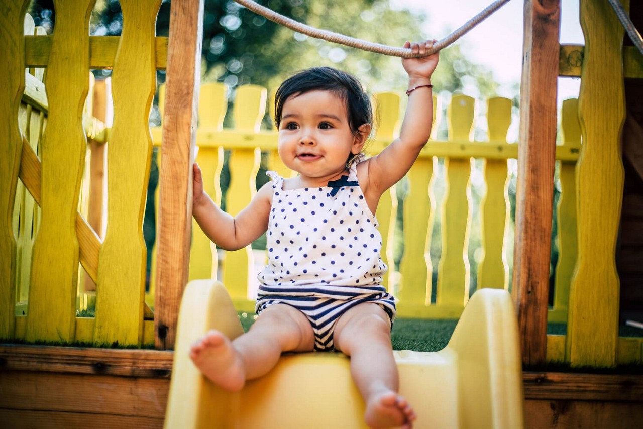 Un jeune enfant vetu d une tenue a pois est assis sur un toboggan jaune vif dans une aire de jeux exterieure. Le cadre presente des structures en bois et une cl�ture jaune vif, avec de la verdure visible en arriere-plan. La scene transmet une ambiance ludique et joyeuse, ideale pour les themes de la famille ou de l enfance.