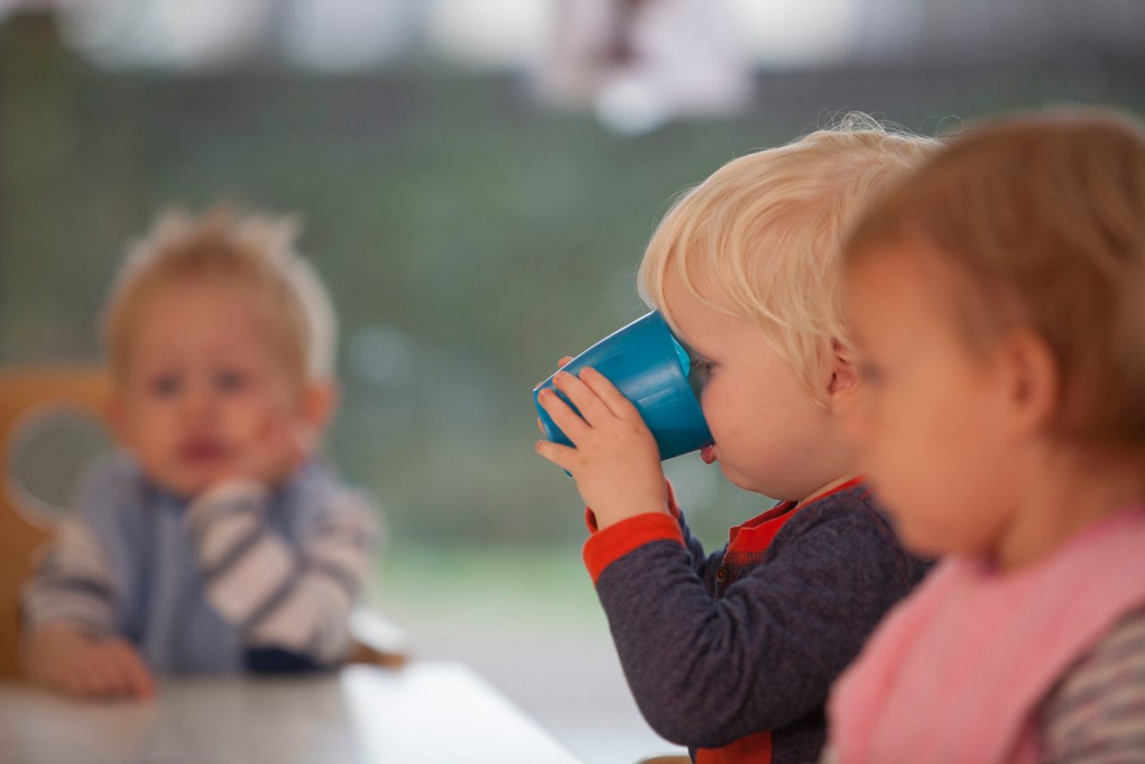 Peuter drinkt uit een beker aan tafel tijdens een gezamenlijke maaltijd, wat zelfstandig drinken en de ontwikkeling van dagelijkse vaardigheden ondersteunt.