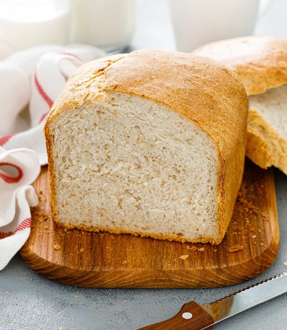 A golden-brown loaf of bread sits on a wooden cutting board, showcasing its soft texture and crusty exterior. The setting includes a knife, a striped cloth, and glasses of milk in the background, suggesting a breakfast or snack scene. The bread is sliced, revealing its fluffy interior.