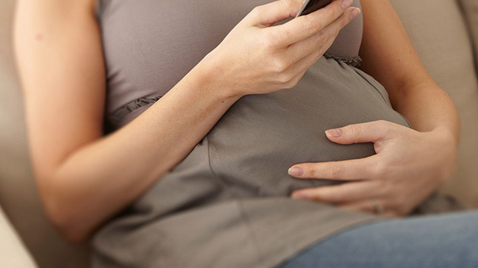 Mujer embarazada sentada en el sofá usando su teléfono, destacando el cuidado prenatal y el seguimiento del embarazo, contenido de Nutricia Club (Guía para embarazadas)