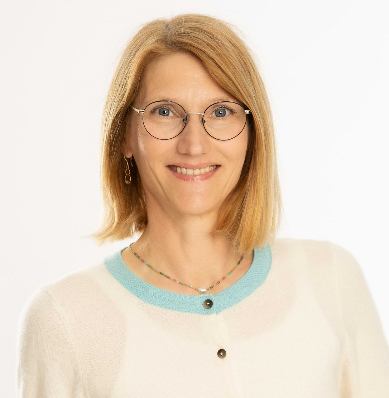 Eine Frau mit Brille und schulterlangem blondem Haar trägt eine weiße Strickjacke vor hellem Hintergrund.